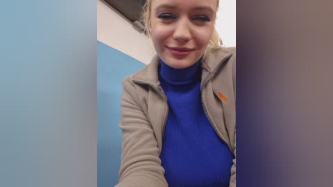 Video of a smiling, light-skinned woman with blonde hair, wearing a blue turtleneck under a beige jacket, standing in a narrow, blue and white hallway.