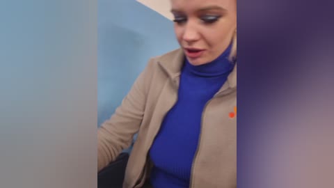 Video of a light-skinned woman with blue eyes, wearing a blue turtleneck, beige jacket, and a small orange pin, standing against a light blue and purple wall, with a blurred background.