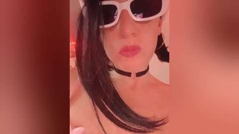 Video of a young woman with fair skin, black hair, and white sunglasses, wearing a black choker and pink lipstick, kissing. The background is blurred, with a warm, reddish-orange tint.