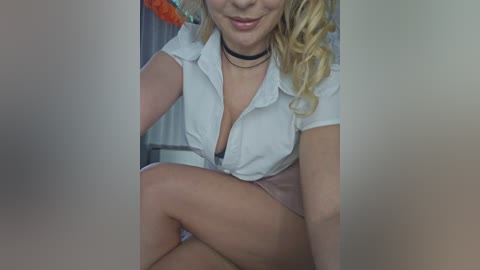 Video of a smiling, blonde woman with wavy hair, wearing a white blouse, black choker, and pink shorts, sitting with legs crossed, indoors.