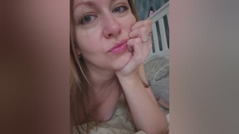 Video of a Caucasian woman with light skin and long blonde hair, resting her chin on her hand, wearing a pink lipstick. Background shows a white crib and a soft blanket.