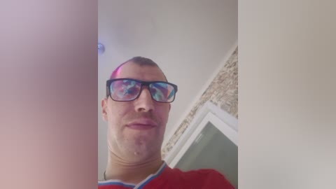 Video of a bald man with a red mohawk, wearing sunglasses and a red shirt, standing in front of a beige wall with a textured brick section.