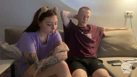 Video of a tattooed woman in a purple shirt and a bald man in a maroon shirt sitting on a beige couch, both looking glum.