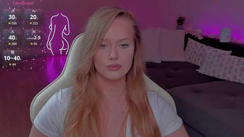 Video of a blonde woman with fair skin and long hair, sitting in a white chair, in a dimly lit bedroom with purple lighting, displaying a virtual avatar on a screen.