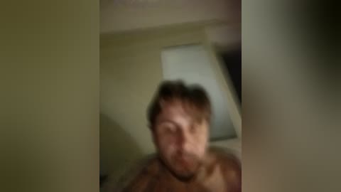 Blurry video of a shirtless man with messy hair, likely in his late 20s or early 30s, standing in a dimly lit room with a window and a light fixture visible in the background.
