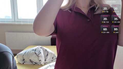 Video of a woman in a maroon polo shirt, holding her hair, standing near a bed with a white pillow and duvet. A digital overlay shows health stats and location data.