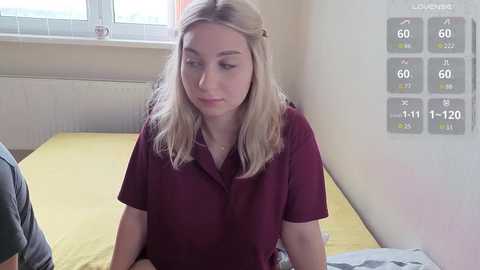 Video of a young woman with long blonde hair, wearing a maroon polo shirt, sitting on a yellow bedspread in a well-lit bedroom. A temperature gauge with 60 degrees Fahrenheit is visible on the wall.