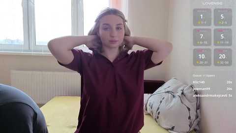 Video of a young woman with light brown hair, wearing a burgundy shirt, standing in a bright, simple bedroom with a yellow bedspread, radiator, and window.