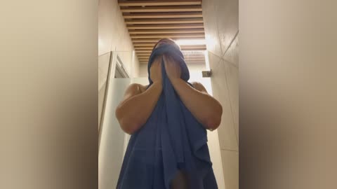 A video of a person, likely male, wearing a blue towel, covering their face, in a modern, beige-tiled hallway with wooden beams on the ceiling.