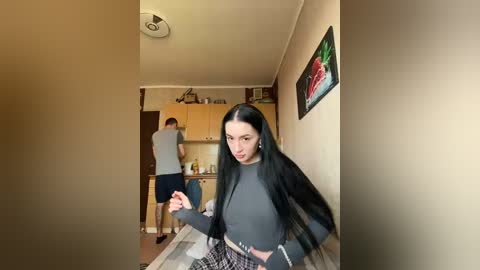 A video of a young woman with long black hair, wearing a grey top and plaid skirt, walking down a narrow hallway, passing a man in a grey shirt and dark pants.