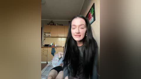 A video of a young woman with long black hair, pale skin, and a slight smile, sitting in a small kitchen with light wooden cabinets, a tiled backsplash, and a colorful floral painting on the wall.