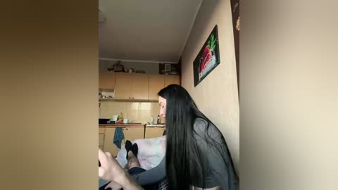 A video shows a young woman with long black hair, wearing a gray shirt, sitting on a bed in a dimly lit, small room with beige walls. A kitchen with wooden cabinets and a colorful painting hangs on the wall behind her.