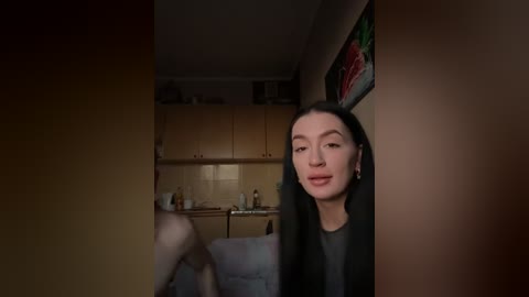 Video of a young woman with fair skin and straight black hair, wearing a black coat, standing in a dimly lit kitchen with wooden cabinets and a small table.