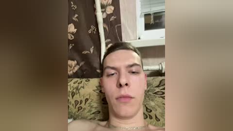 Video of a young, fair-skinned man with short brown hair, lying on a patterned couch with floral curtains behind him. He has a neutral expression, and the room has a warm, cozy ambiance.