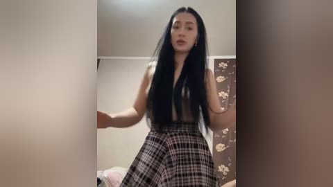 Video of an East Asian woman with long black hair, wearing a black sleeveless top and a high-waisted, checkered skirt, standing in a room with beige walls and floral-patterned wallpaper.