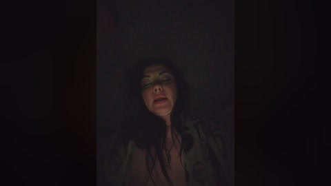 A dimly lit video of a woman with long, dark hair and light skin, wearing a gray shirt, lying on a dark surface with a distressed expression, eyes closed, mouth slightly open.