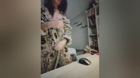 Video of a woman with curly black hair in a leopard-print robe, standing in a dimly lit room with shelves of books, a desk, and a black mouse.