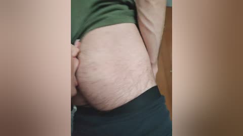 A video of a person's lower body, showing pale skin with stretch marks and cellulite, wearing dark pants, partially lifted green shirt, revealing a belly. Background shows a wooden door.