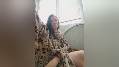 Video of a middle-aged woman with dark hair, wearing a patterned robe, sitting on a bed with a window in the background, partially obscured by a large object.