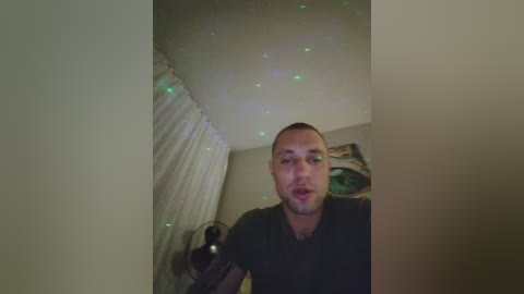 A video of a man with short hair, wearing a black shirt, standing in a dimly lit room with white curtains and a green wall poster.