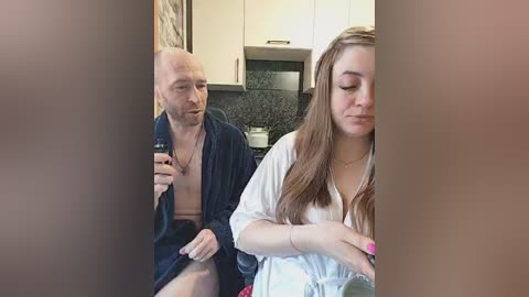 A candid video shows a bald, shirtless man in a robe, holding a bottle, and a young woman with long brown hair, wearing a white robe, seated in a modern kitchen with white cabinets and a black countertop.