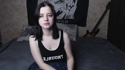 Video of a young woman with shoulder-length black hair, wearing a black tank top with white text, sitting on a bed with a guitar and a tapestry in the background.