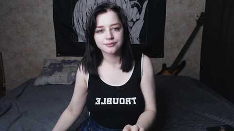 A video of a young woman with pale skin and shoulder-length black hair, wearing a black \"THORN\" tank top and blue jeans, seated on a bed with a guitar and tapestry in the background.