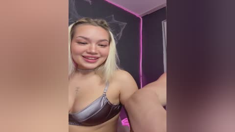 Video of a smiling, fair-skinned woman with blonde pigtails in a silver bra, standing in front of a dark wall with purple lighting.