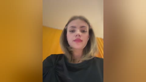 A video of a woman with shoulder-length blonde hair, wearing a black blouse, standing in a room with mustard-yellow walls. She has a neutral expression.