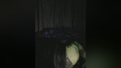 A dimly lit video shows a person, partially obscured, lying on the ground next to a dark, textured wall, possibly in a forest or urban alley. The individual appears to be in distress, with a green and black backpack partially visible.