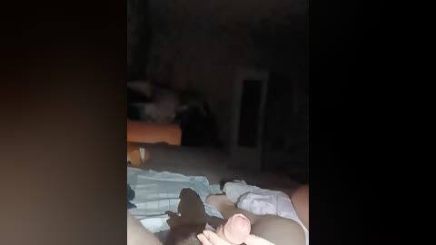 A blurry video shows a woman lying on a bed, partially covered by white sheets, in a dimly lit, cluttered bedroom.