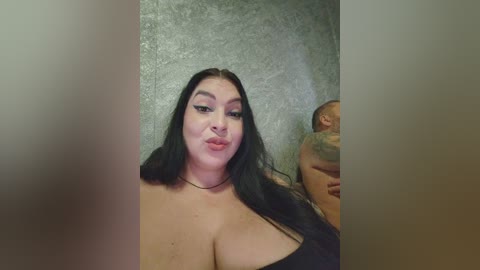 Video of a plus-sized woman with long black hair and fair skin, wearing a black top, posing in front of a textured, grey wall.