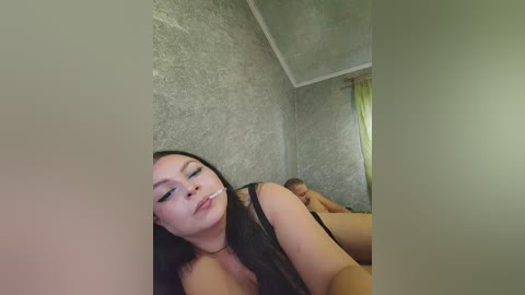 A video shows a nude woman with long dark hair, resting on a bed, her head slightly tilted back, with a cigarette in her mouth. The room has gray textured walls and a yellow curtain.