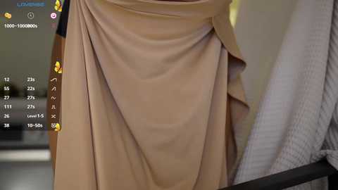A close-up video of a person's torso in a beige dress, with a blurred background featuring a grey textured blanket and a digital interface displaying numerical data and emojis.