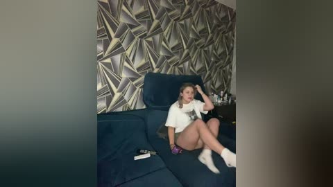A video shows a young woman with blonde hair, wearing a white t-shirt and shorts, reclining on a dark blue sofa. The background features geometric, gold-patterned wallpaper.