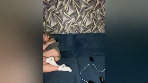 Video of a woman in a gray sweater and white socks, lying on a dark blue couch, with geometric wallpaper behind her, and scattered electronics on the floor.