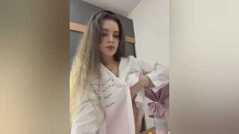 Video of a young woman with long, straight brown hair, fair skin, wearing a white robe with black writing, and a pink scarf, standing in a modern, minimalist room with beige walls.