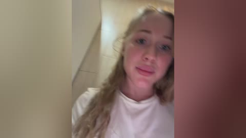 Video of a young Caucasian woman with long blonde hair, wearing a white shirt, standing in a beige-tiled bathroom. She has a neutral expression.