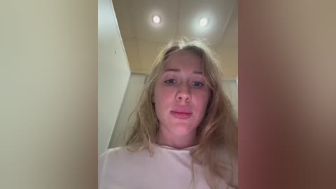 Video of a young Caucasian woman with long, wavy blonde hair, wearing a white shirt, standing in a beige-walled hallway with recessed ceiling lights.