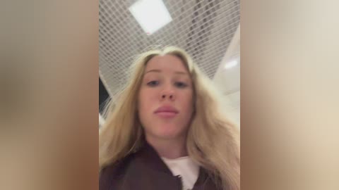 Video of a young woman with long, blonde hair, wearing a black jacket over a white shirt, standing in a modern, industrial-looking room with a grid-patterned ceiling and fluorescent lighting.