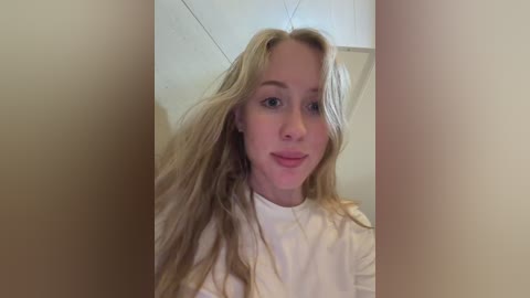 Video of a young woman with long, wavy blonde hair, wearing a white t-shirt, standing in a dimly lit room with beige walls and a white door.