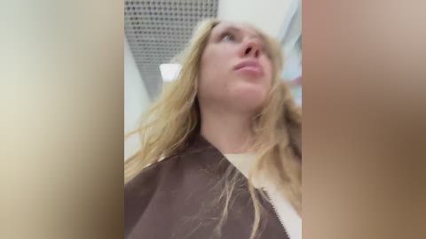 Video of a blonde woman with long hair, wearing a brown cape, looking up with a neutral expression. Background shows a ceiling with a grid pattern.