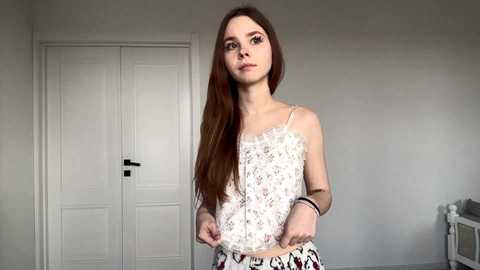 A young woman with long, straight brown hair stands in a minimalist room, wearing a white lace camisole and patterned pants. The background features a white door and a white wall.