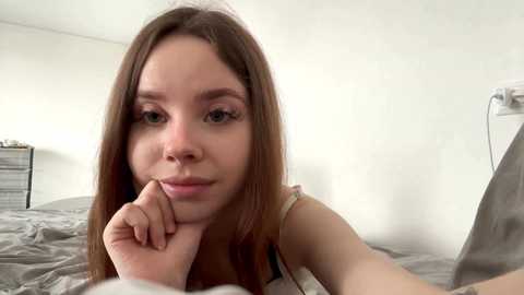 A video of a young Caucasian woman with long brown hair, light skin, and a slender build, lying on a bed with gray sheets. She has a small nose piercing and wears minimal makeup. The room is sparsely decorated with a nightstand and a white wall.