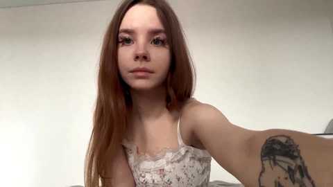 A young Caucasian woman with long, straight brown hair, wearing a white lace camisole, takes a selfie indoors. Her expression is neutral, and she has a small tattoo on her right forearm.