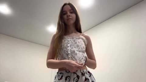 Video of a young, light-skinned woman with long, straight brown hair, wearing a white floral sundress, holding a small, white ceramic mug. The background features a simple, off-white room with recessed ceiling lights.
