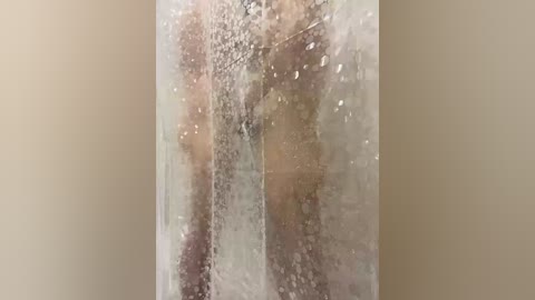 Video of a nude person standing in a fogged-up shower stall, with water droplets on the glass, creating a translucent effect. The background is a plain beige wall.