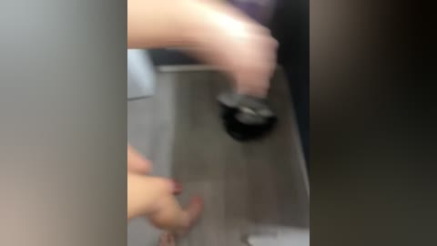 A blurry video shows a person's hand, wearing black nail polish, holding a black object, possibly a phone or tablet, against a grey tiled wall. The image is slightly out of focus.