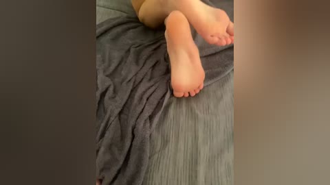 A video of a pair of light-skinned feet, crossed at the ankles, resting on a dark gray blanket. The background is a blurred, neutral-toned wall.