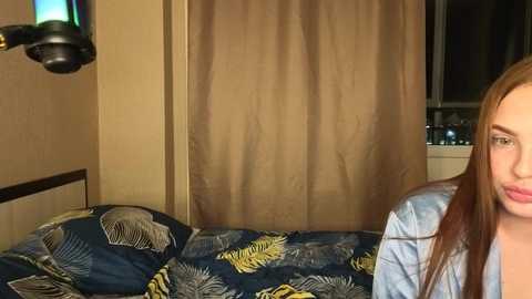 Video of a young woman with long, light brown hair, wearing a blue hoodie, sitting on a bed with a floral-patterned duvet in a beige room with a brown curtain and a window.